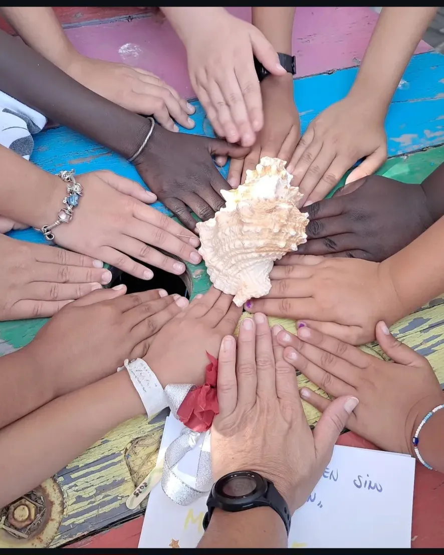 Manos de un grupo de niños alrededor de una caracola sobre una mesa pintada de colores